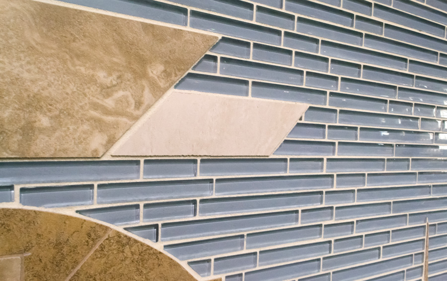 Close-up of diagonal blue glass mosaic brick pattern meeting stone-look ceramic tile fields in tans and creams