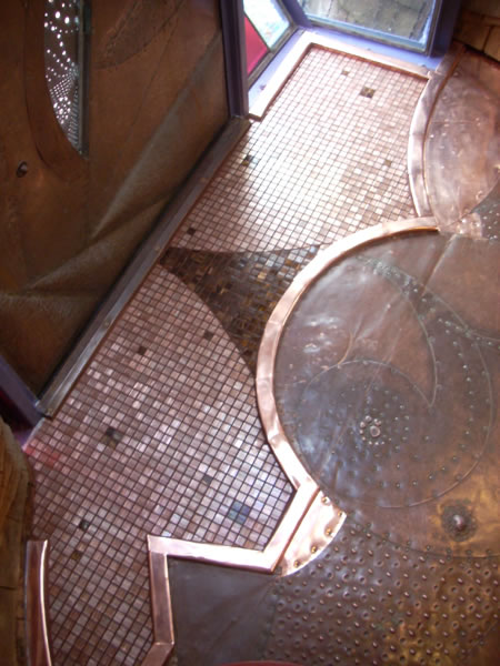 Detail of a copper-clad raised disc rising from copper-toned mosaic tile floor with darker glass tile triangular accent inset