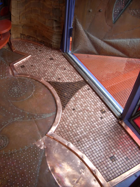 Overhead view of an entryway floor — copper mosaic tile with curved organic copper-trimmed shapes, decorative dot patterns, and copper-mesh doormat grate