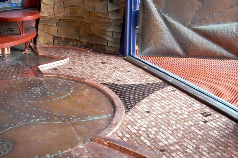 Detail of copper-toned mosaic tile floor with copper trim curling into organic shapes, meeting a textured shingled wood wall and copper-mesh threshold
