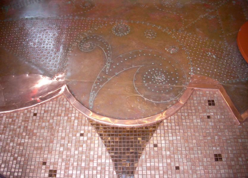 Detail of a copper-clad floor panel with etched vine and leaf patterns over a dotted rivet ground, meeting copper-toned mosaic tile with darker accent squares