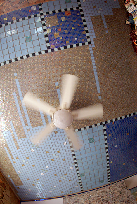 Looking up at a mosaic-tiled ceiling with a white ceiling fan in motion, blue and beige iridescent glass tile arranged in checkered patterns