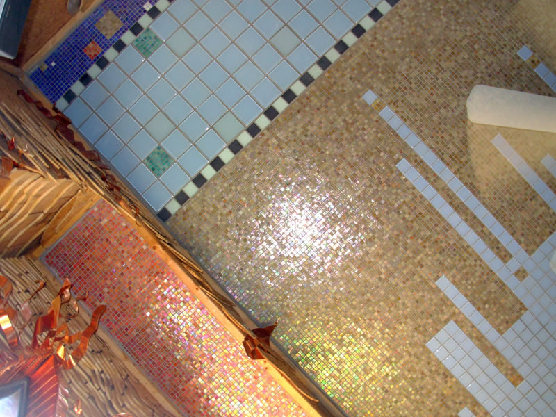 Overhead view of a mosaic-tiled surface: tan iridescent glass tile field intersected with white tile stripes and bordered by blue panels