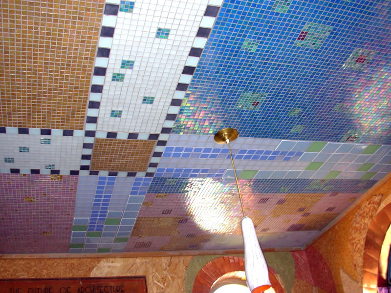 Mosaic-tiled ceiling with brass pendant fixture — tan striped, deep blue, and pink iridescent panels above a wall inscribed "The Future of Architecture"