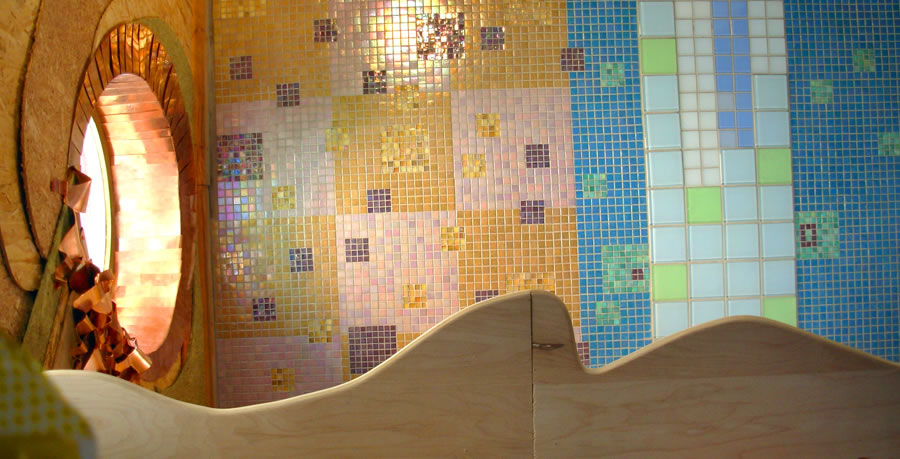 Round wooden alcove with copper-tile interior set into a wall of gold and blue iridescent glass mosaic, curved plywood shelf in foreground
