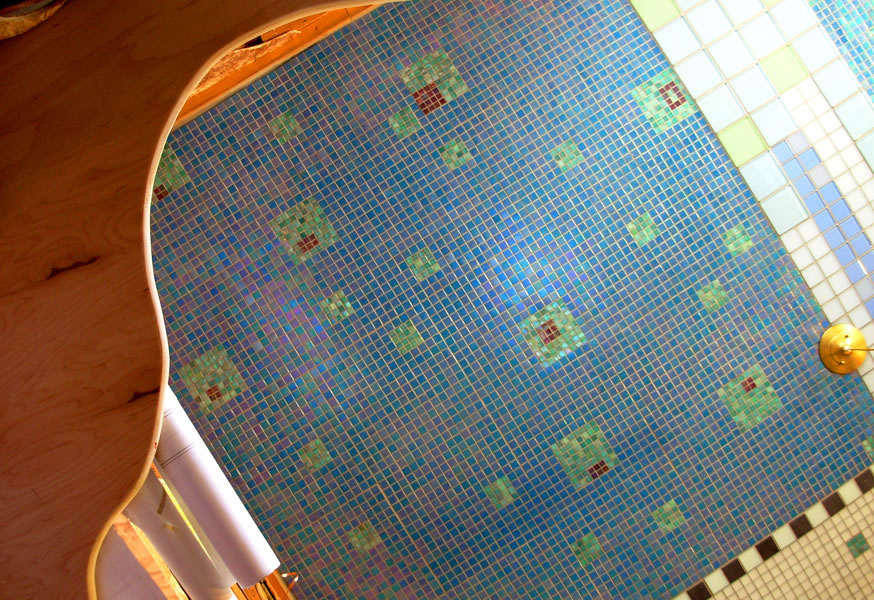 Iridescent blue mosaic tile surface with scattered green and reddish-brown square accent tiles, curved plywood edge in foreground