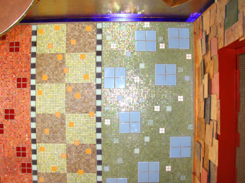 Vertical mosaic panels of olive green and pink iridescent glass tile with square accent tiles meeting a rough stone wall