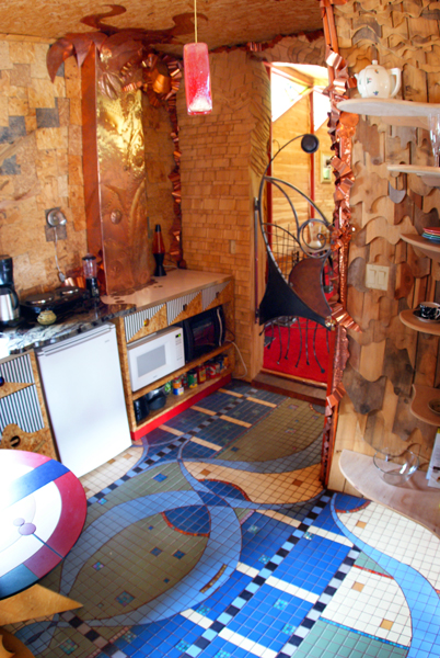 Wahopi kitchen with hammered copper backsplash and pendant light, copper-clad lower cabinets, sculpted bookshelf wall divider, and an intricate mosaic floor of curved organic shapes in blue, cream, and sage with checker pattern bands