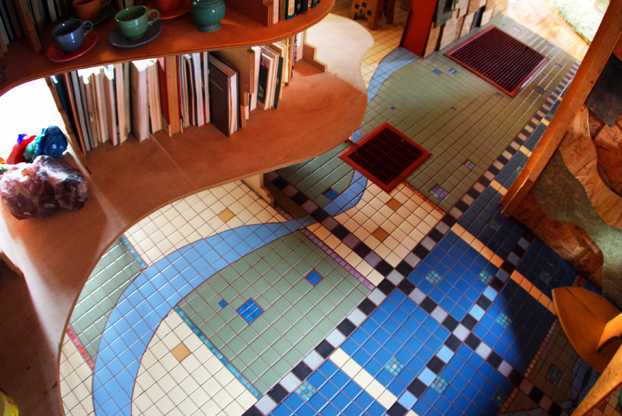 Overhead view of a mosaic-tiled kitchen floor — sweeping curves of blue, sage, and cream tile in interlocking circular patterns separated by grid registers and checker pattern bands, beside a built-in curved bookshelf