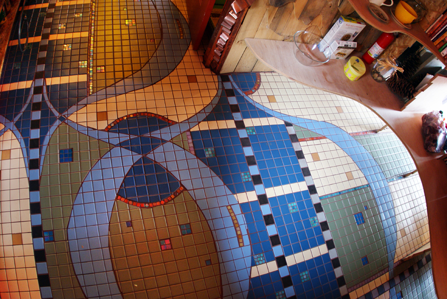 Detail of the Wahopi kitchen mosaic floor — interlocking ovoid shapes in blue, sage, and gold with white grid registers and small accent tiles, meeting wood plank flooring at the edges
