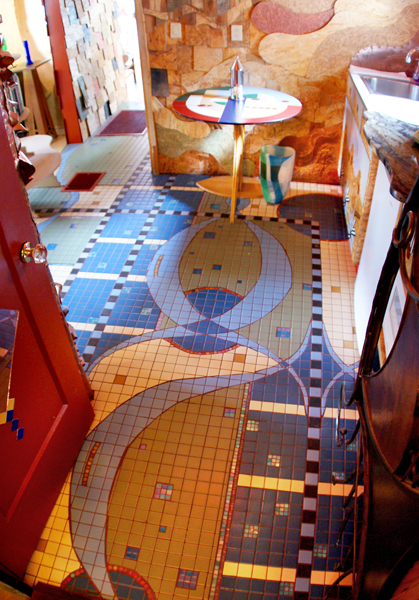 Wahopi kitchen entry — door opening to a mosaic floor of curved organic shapes in blue, cream, and sage with red accent dots, sculpted stone wall, and a hand-painted plate on a side table