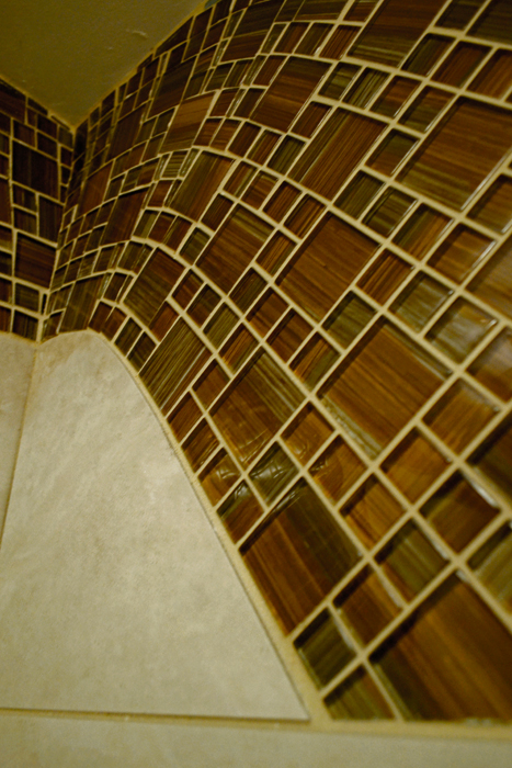Detail of brown striped glass mosaic wave wrapping around a shower corner, meeting beige porcelain tile with a tapered transition