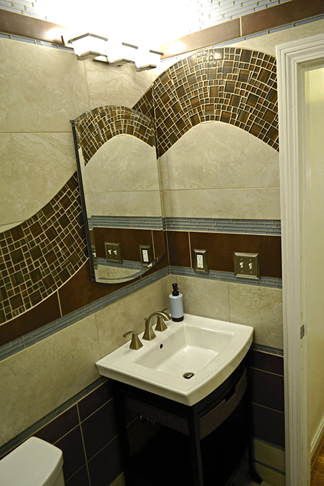 Bathroom vanity area with brown glass mosaic wave arcing over the mirror, beige porcelain tile walls, pedestal sink, and brushed nickel faucet