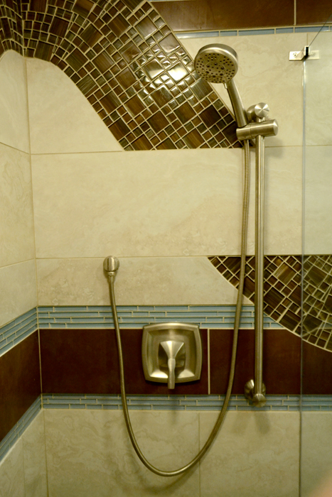 Shower fixture detail — handheld showerhead on a slide bar with the curved brown glass mosaic wave arcing overhead and pale blue glass tile banding below