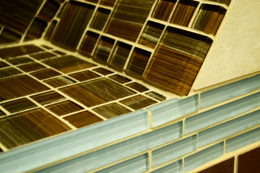 Detail of a beige porcelain tile bench seat meeting a brown glass mosaic backsplash with a pale blue glass linear tile transition band