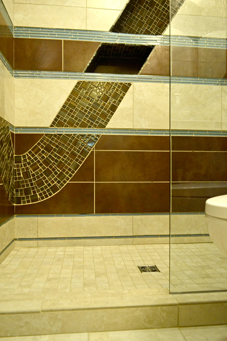 Inside shower view — brown glass mosaic wave curving across beige porcelain tile walls, dark brown porcelain horizontal bands, pale blue glass trim, and glass enclosure