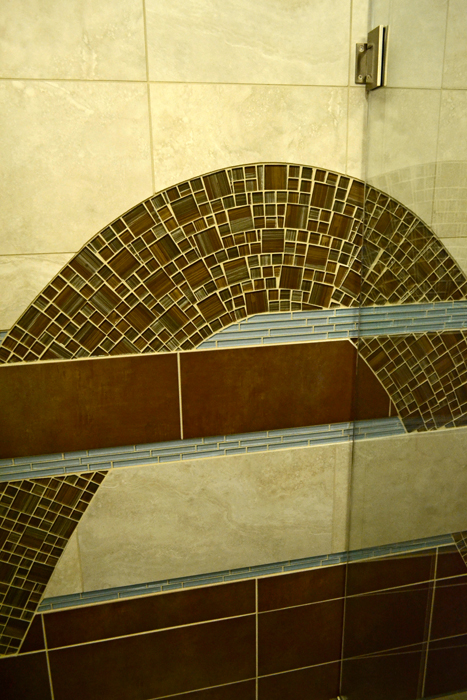 Detail of the arched curve of the brown glass mosaic wave meeting horizontal bands of beige and dark brown porcelain tile with pale blue glass trim banding